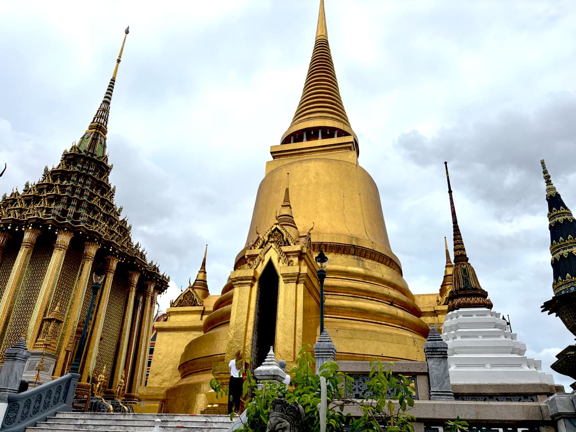Golden spires and ornate temples of the Grand Palace in Bangkok, under a cloudy sky.