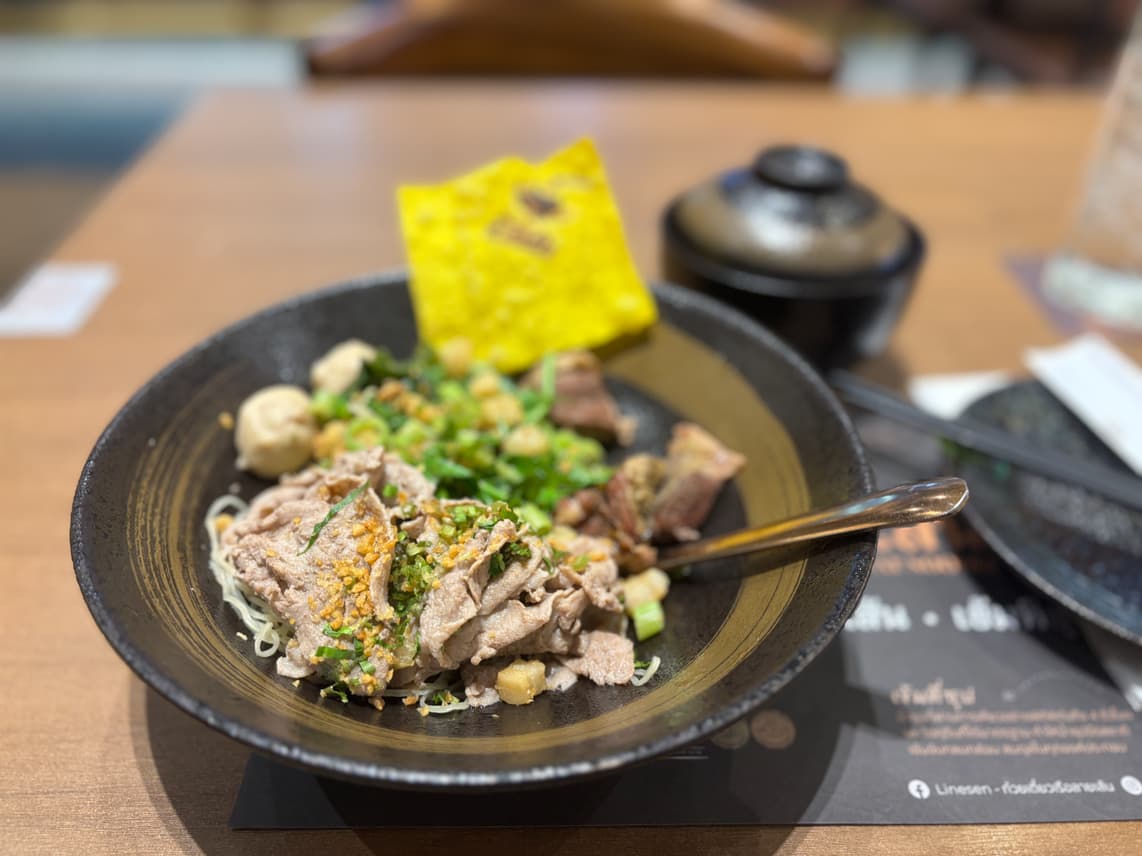 Thai boat noodles with beef, meatballs, herbs, and a crispy wonton, served in a black bowl with soup on the side.