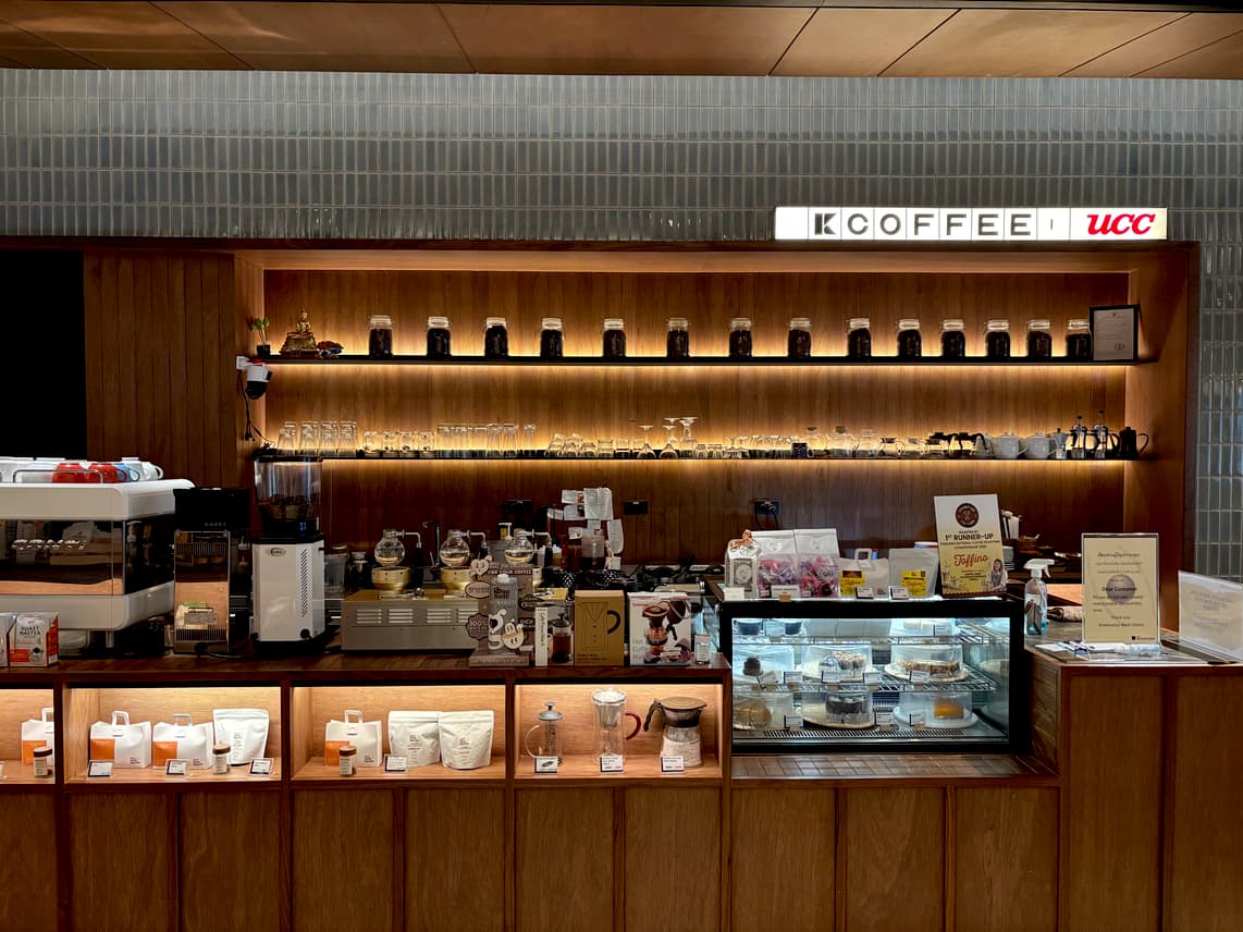 A bright coffee shop counter with wooden shelves displaying coffee beans, brewing equipment, and pastries, under “K COFFEE” and “UCC” signs.