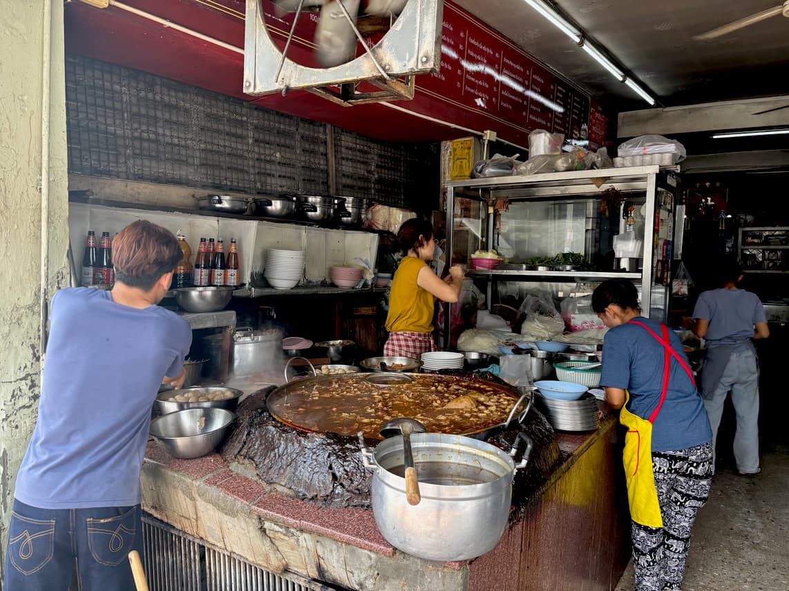 A bustling, open-air Thai kitchen where several cooks work around a large, simmering cauldron of stew.