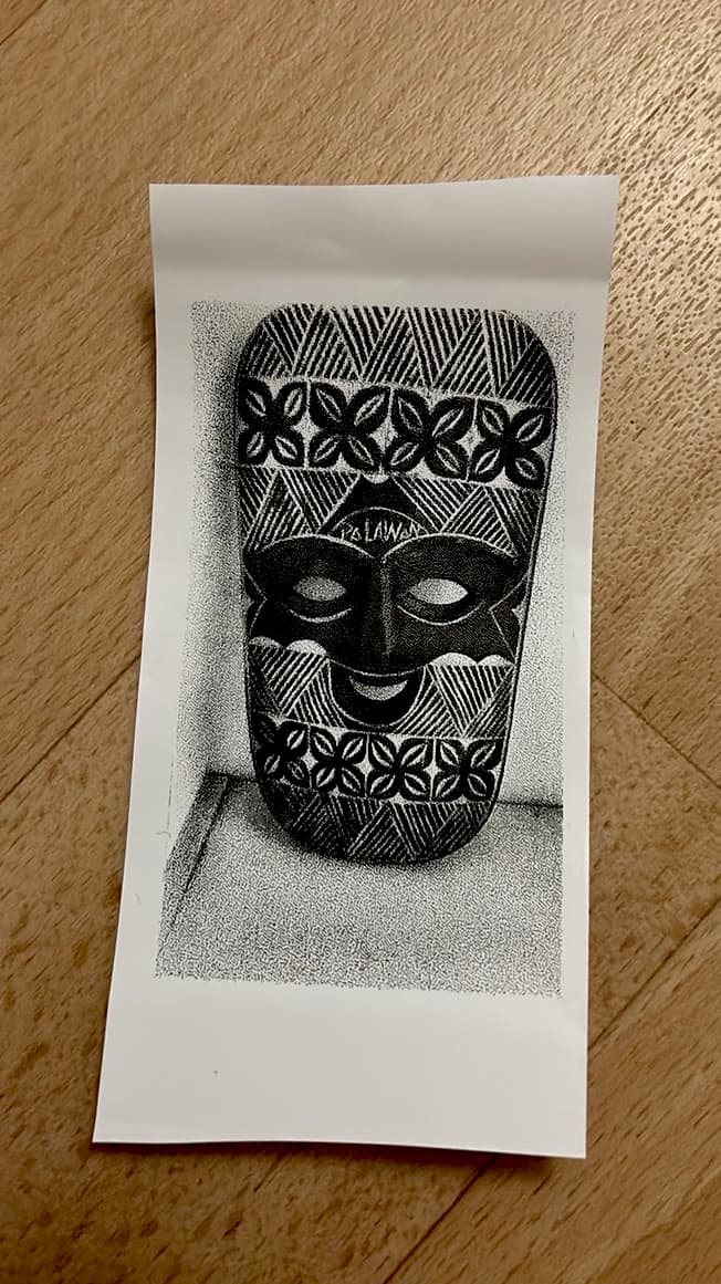 A black and white printout on thermal paper of a stippled drawing of a decorative, patterned mask, resting on a wooden surface.