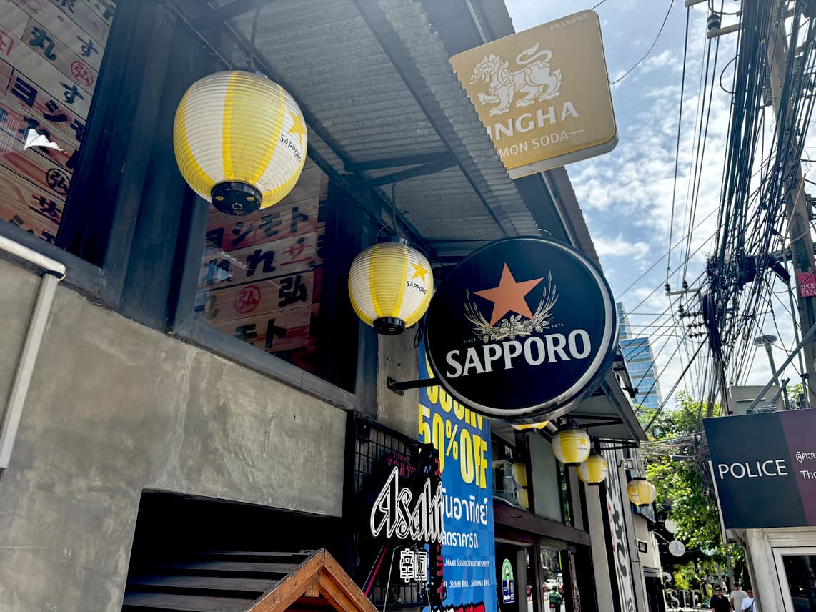 Japanese-style restaurant exterior in Bangkok with Sapporo and Singha signs, lanterns, and promotional banners on a sunny street.