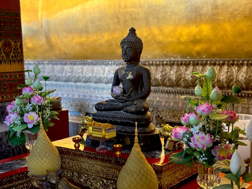  A small black Buddha statue in a meditative pose, holding a pink lotus flower, placed on an ornate altar with golden decorations, artificial lotus arrangements, and traditional Thai offerings inside a richly decorated temple interior.