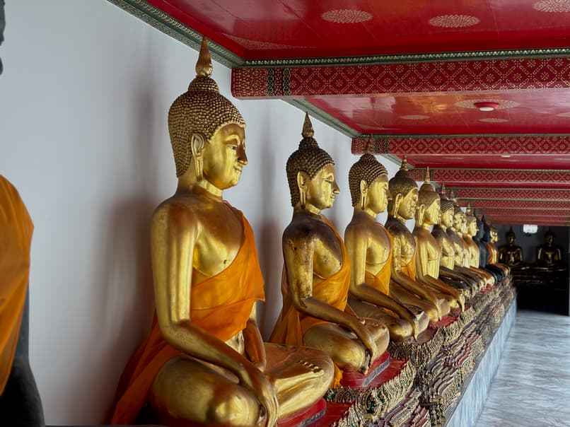 A long row of golden Buddha statues, draped in orange robes, sit in meditation postures along a white wall under a red ceiling at Wat Pho.