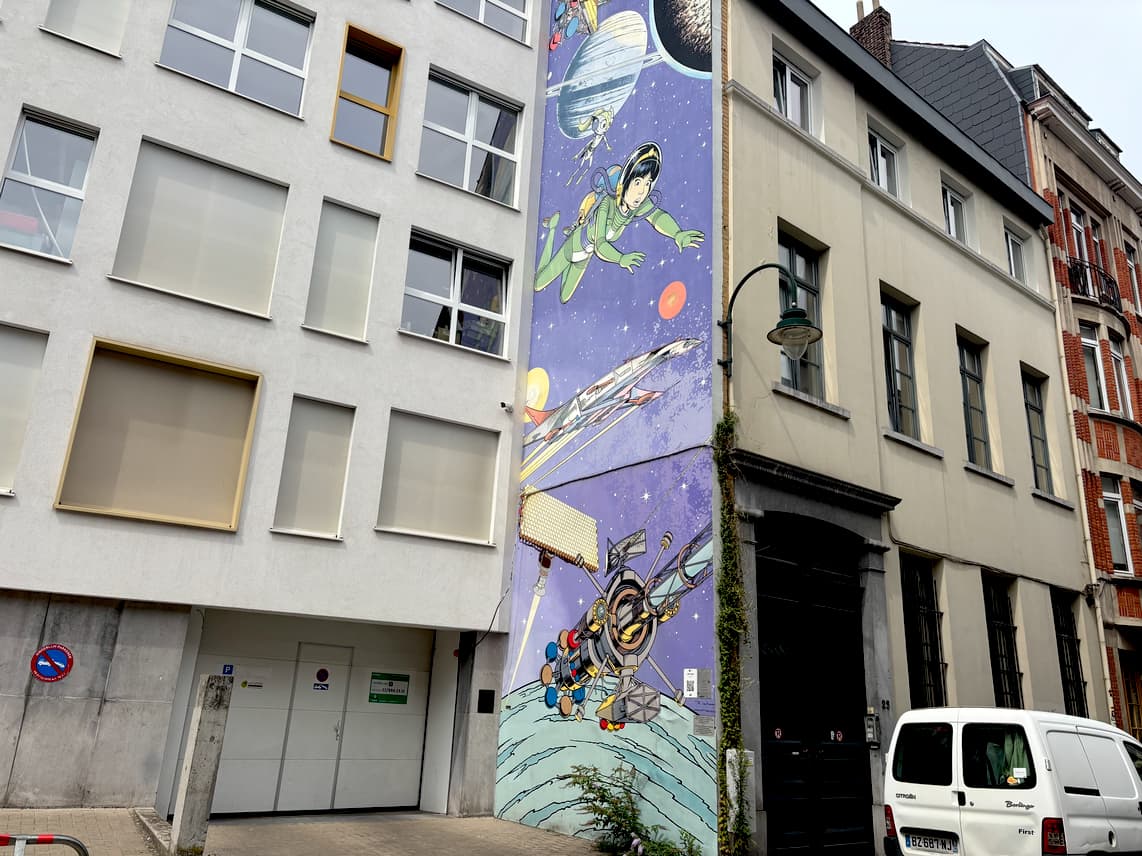 A tall mural on the side of a building depicts a cartoon woman in a green flight suit flying over a futuristic cityscape with spaceships and planets.